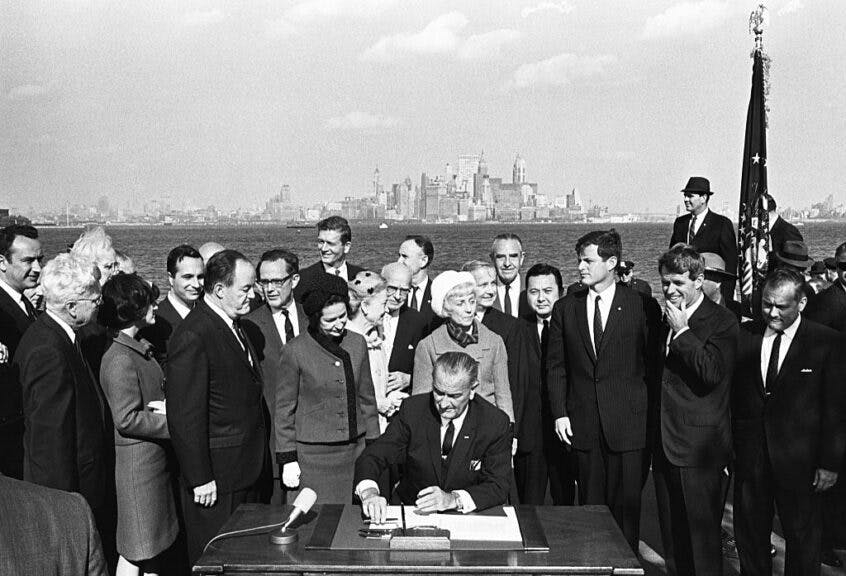 President Lyndon B. Johnson signs the Immigration Bill of 1965 on Liberty Island in New York Harbor with a view of the New York City skyline in the background. Next to the president on his right are First Lady Lady Bird Johnson and Vice President Hubert Humphrey. To the president's left are Senator Edward Kennedy (third from right) and Robert Kennedy (second from right). (Photo by © CORBIS/Corbis via Getty Images)