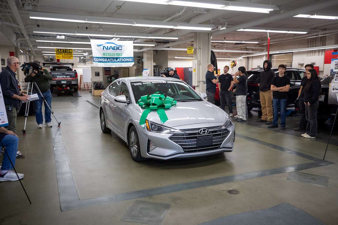 A silver Hyundai sedan adorned with a large green bow is showcased in a garage, surrounded by people and media, with a banner celebrating 
