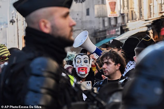 A person wears a Halloween mask while a man speaks on a megaphone behind him on the streets of Paris