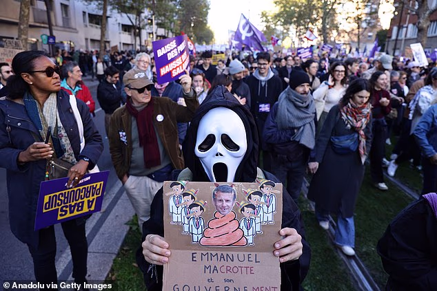 A protester wearing a Scream mask holds a placard depicting French president Emmanuel Macron